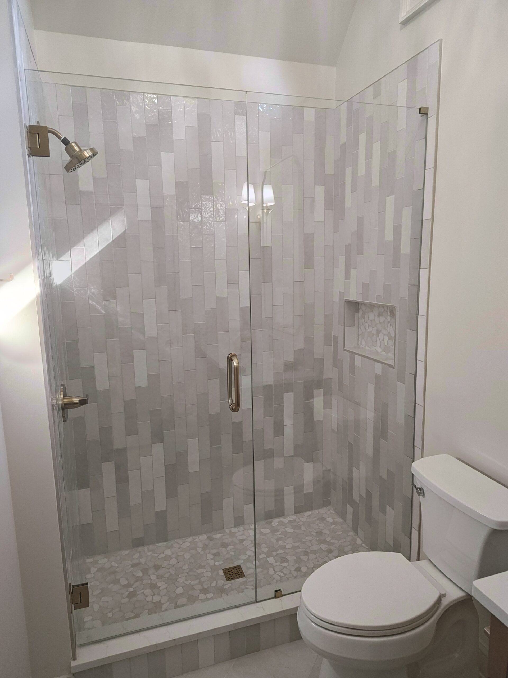 Glass-walled shower with gray rectangular tile walls, a pebble-textured floor, and a toilet beside it in a modern bathroom.
