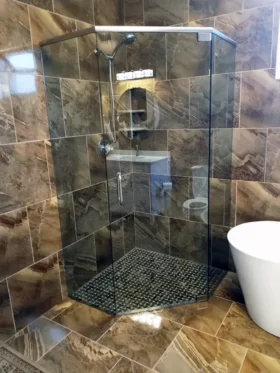 Neo-angle frameless shower with a brushed nickel support header, clear glass, brown marble-look tile, dark mosaic floor, corner shelf, and freestanding tub.